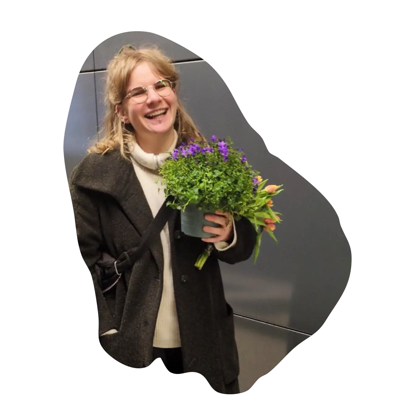 Porträt von Julia. Sie steht lachend mit einem Blumenstrauß vor einer grauen Wand.